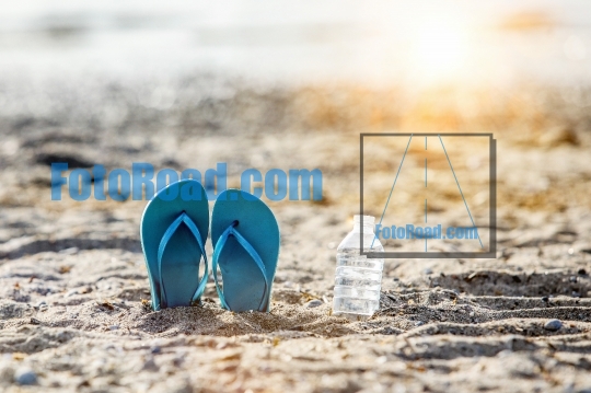 Flip flops on beach sand with water in plastic bottle and sunbea